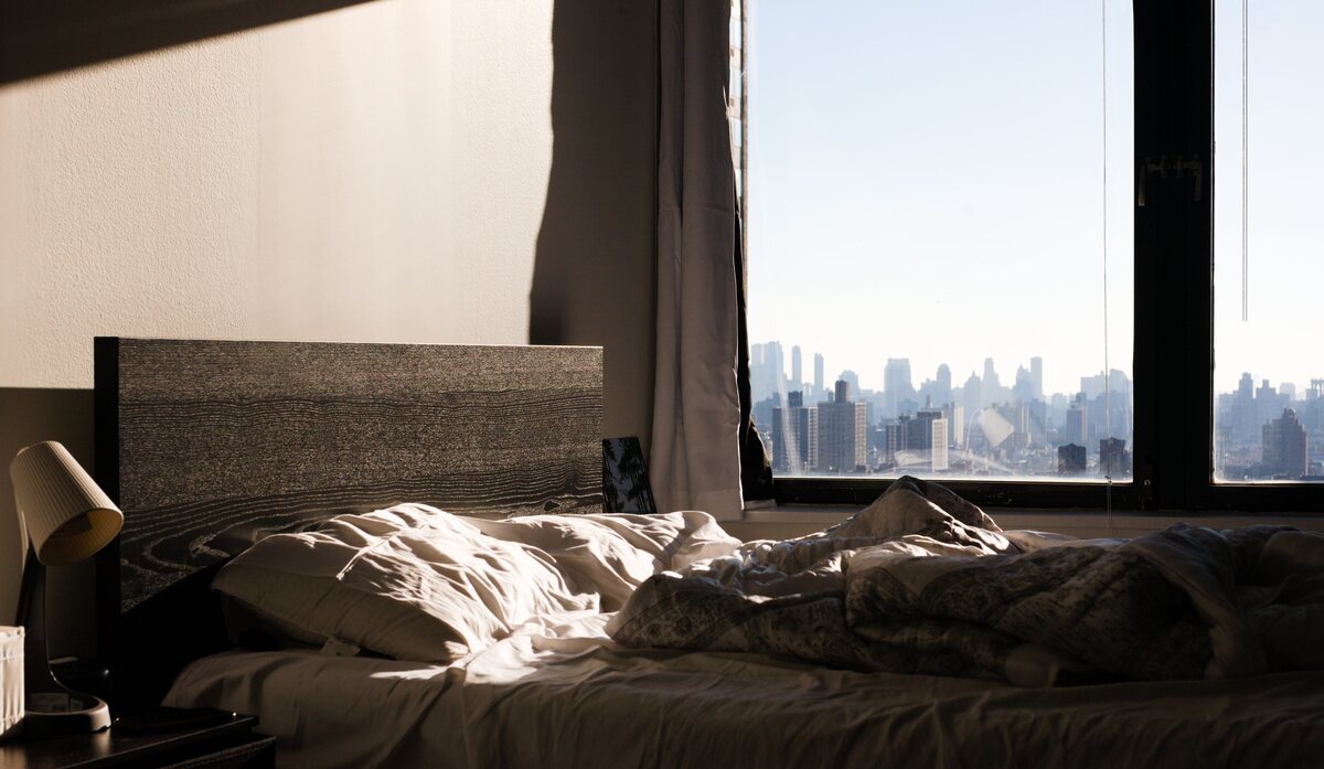An unmade bed by a window at dawn with a city skyline